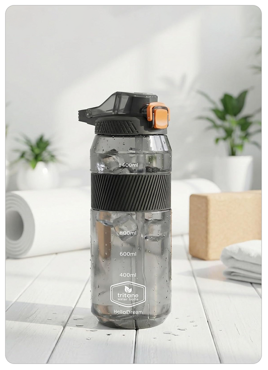 A translucent gray Tritane water bottle with a black flip-top lid and orange accent, standing on a white wooden surface with a softly blurred home background.