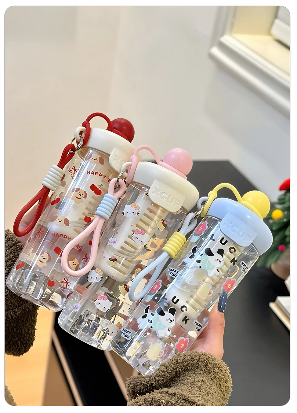 clear plastic water bottles with cute cartoon prints and colorful lids are held in a hand. The bottles have matching wrist straps and are decorated with characters and text including 'LUCK' and 'HAPPY'.
