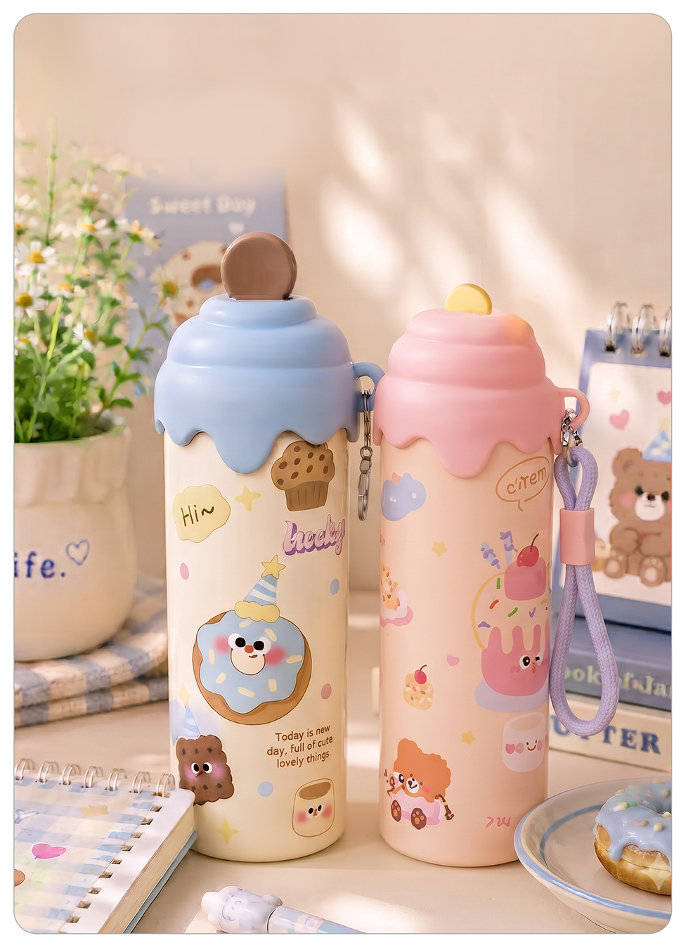 cute cartoon water bottles styled on a tabletop with notebooks, a plant, and a plate in the background. One bottle is cream with a light blue frosting-like top and donut illustrations, and the other is pale pink with a pink frosting-like top and cake illustrations.