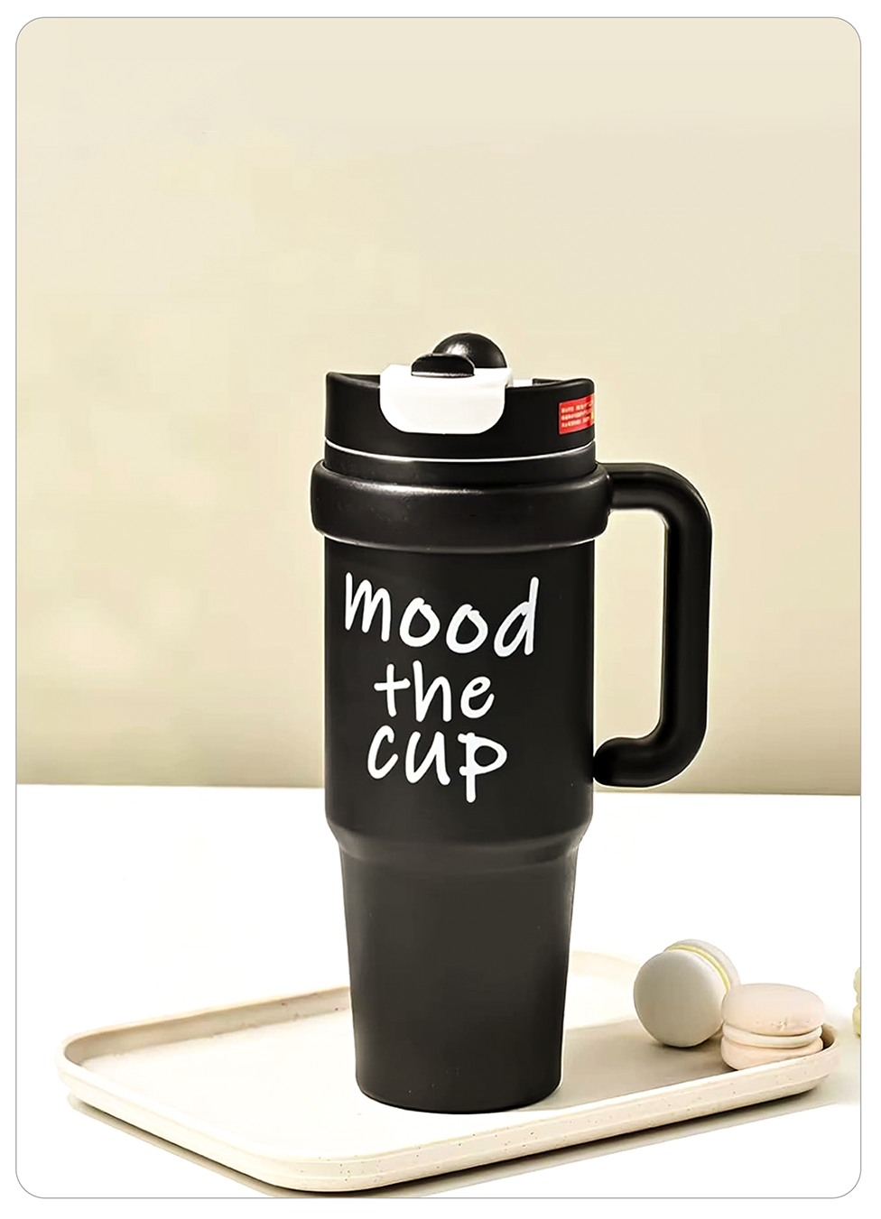 A black insulated travel tumbler with a flip-top lid and handle, printed with the phrase 'mood the cup' in white script, displayed on a cream tray with two macarons against a beige background.