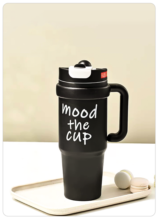 A black insulated travel tumbler with a flip-top lid and handle, printed with the phrase 'mood the cup' in white script, displayed on a cream tray with two macarons against a beige background.