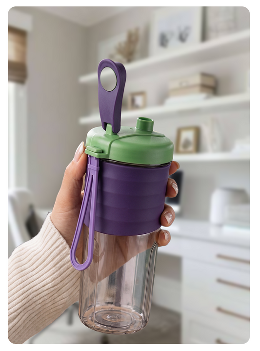 A hand holding a clear plastic shaker bottle with a purple silicone sleeve, green lid, and purple carrying loop in a bright, modern room setting.