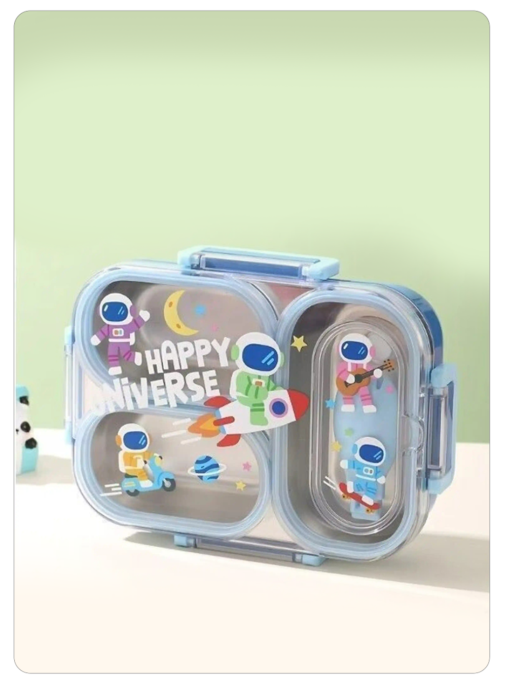 A light blue double-compartment lunch box with clear plastic sides, decorated with colorful astronaut stickers and a 'HAPPY UNIVERSE' graphic, set against a soft green and cream background.