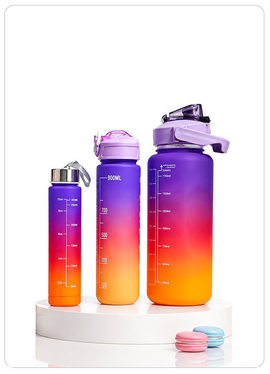 Three reusable water bottles in graduated sizes with a purple-to-orange gradient design, displayed on a white pedestal with two macarons in the foreground against a white background.